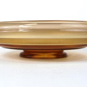 Könnte beinhalten: Eine Vintage-Schale mit Fuß aus bernsteinfarbenem Glas. Die Schale hat ein breites, flaches Design mit einem abgerundeten Rand und einem abgestuften Sockel. Das Glas hat eine glatte, glänzende Oberfläche und die Farbe variiert von hell- bis dunkelbernsteinfarben.