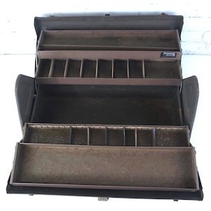 Puede incluir: Una caja de herramientas de metal de color marrón oscuro con múltiples bandejas de almacenamiento escalonadas. El interior cuenta con varios compartimentos para organizar y una pequeña etiqueta negra con el texto blanco Kennedy es visible.