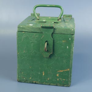 May include: A vintage green wooden box with a metal handle and latch. The box has a rectangular shape and shows signs of wear, with visible paint chipping. The metal hardware has a rustic appearance. The box is likely used for storage.