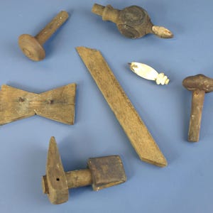 Può includere: Assortimento di antichi utensili e componenti in legno, tra cui un martello, un pezzo a forma di farfallino e varie parti in legno tornito. Gli oggetti sono in tonalità marroni e beige, su sfondo blu.