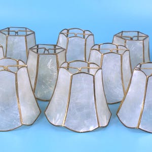 May include: A collection of twelve bell-shaped lamp shades featuring translucent white capiz shell panels framed in thin gold-toned metal. Each shade has a scalloped bottom edge and a wire fitter at the top, arranged in a group on a light blue surface.