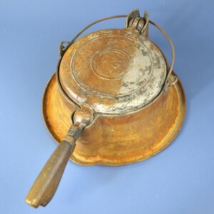 May include: A vintage cast iron waffle iron with a round base and a long handle. The metal surface shows significant rust and oxidation. A circular emblem featuring a profile of a person wearing a headdress is embossed on the top lid.