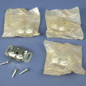 Pode incluir: Conjunto de ferragens com um fecho de porta de armário de metal com dois rolos brancos, três parafusos e três conjuntos de rolos de porta de armário em embalagem de plástico. Os itens são prateados e brancos, sobre um fundo azul.