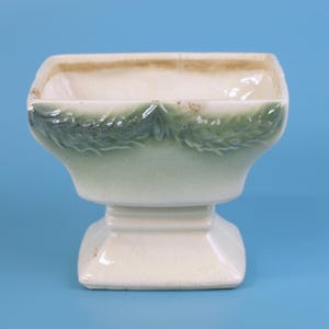 Pode incluir: Um vaso de cerâmica quadrado, branco sujo, com base de pedestal. A borda superior tem uma cor castanha clara, e o vaso é decorado com um padrão de folhas verdes. O vaso está sobre um fundo azul.