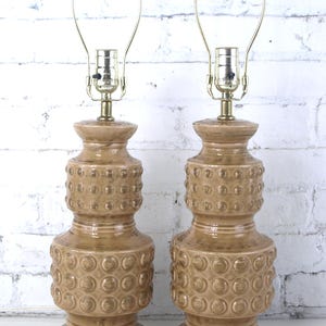 May include: A pair of matching table lamps featuring a tan, glazed ceramic base with a stacked, tiered silhouette. Each base is decorated with a repeating embossed circular pattern and topped with a brass-toned metal harp and light socket assembly.