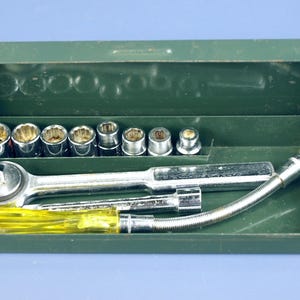 Puede incluir: Una caja de herramientas de metal verde que contiene un juego de vasos cromados, una llave de carraca, una extensión flexible y un destornillador con mango amarillo. Las herramientas están dispuestas ordenadamente dentro de la caja abierta.
