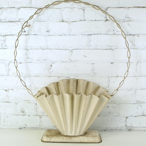 May include: Cream-colored, fan-shaped metal planter with a decorative, circular handle. The planter sits on a rectangular base and is set against a white brick wall. The handle is made of twisted metal.