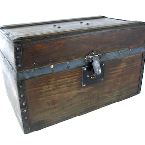 Puede incluir: Un baúl de madera vintage con madera marrón oscuro y detalles metálicos. El baúl tiene forma rectangular con tapa con bisagras y cierre metálico. Las esquinas están reforzadas con tiras y remaches metálicos, lo que le da un aspecto rústico.