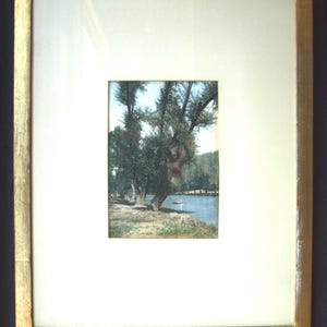 May include: A framed landscape print featuring a serene waterside scene. The artwork depicts trees lining the shore of a calm body of water, with a small boat visible. The frame is gold-toned with a white mat.