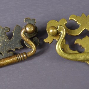 May include: Two ornate brass drawer pulls with intricate designs. One pull is a traditional loop style, while the other has a more modern, curved design.