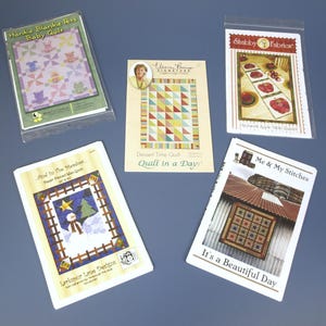 Könnte beinhalten: Fünf verschiedene Quiltmuster in Plastiktüten. Die Muster umfassen "Hankie Blankie Pets Baby Quilt", "Dessert Time Quilt Quilt in a Day", "And to the Meadow Paper Pieced Wall Quilt", "Shabby Fabrics Patchwork Apple Table Runner" und "Me & My Stitches It's a Beautiful Day".