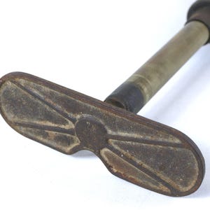 May include: A vintage metal hand pump featuring a weathered, oval-shaped base with a raised, symmetrical geometric pattern. The pump has a cylindrical brass-toned shaft that extends upward, showing signs of oxidation and age-related wear throughout.