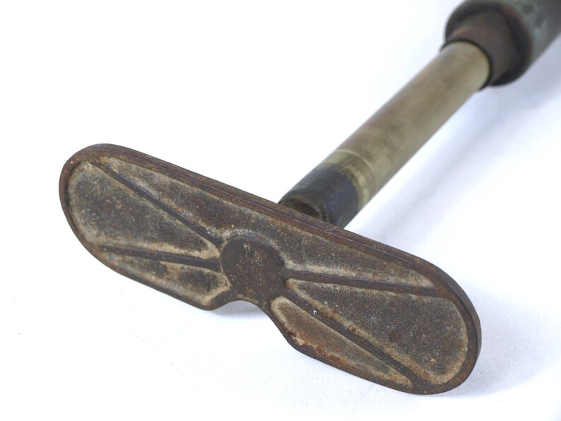 May include: A vintage metal hand pump featuring a weathered, oval-shaped base with a raised, symmetrical geometric pattern. The pump has a cylindrical brass-toned shaft that extends upward, showing signs of oxidation and age-related wear throughout.