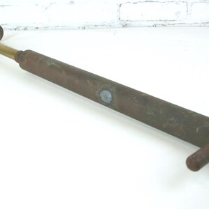 May include: A vintage manual air pump featuring a long, cylindrical, weathered brown metal body with a brass-colored piston rod. It includes a T-shaped handle at one end and a cross-bar base at the other, showing signs of oxidation and age.
