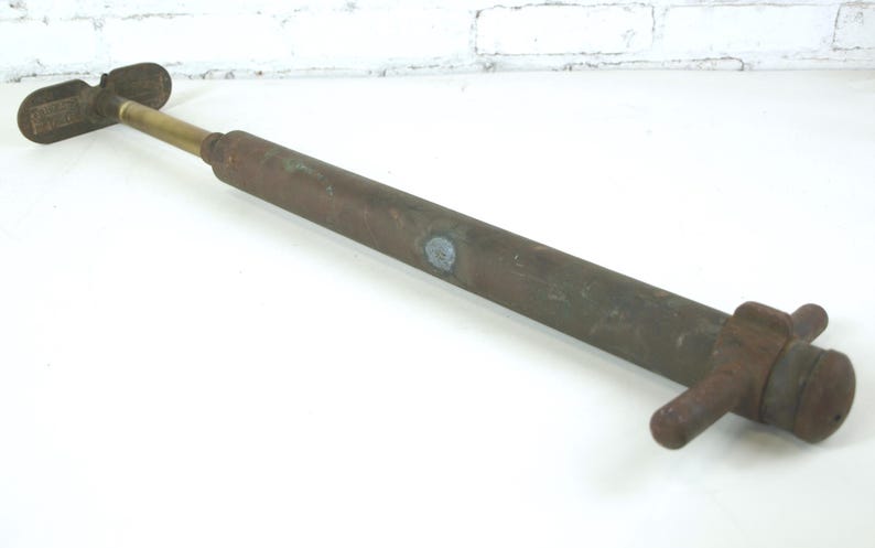 May include: A vintage manual air pump featuring a long, cylindrical, weathered brown metal body with a brass-colored piston rod. It includes a T-shaped handle at one end and a cross-bar base at the other, showing signs of oxidation and age.