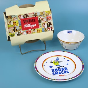 May include: A vintage Kellogg's cereal box with a red "Vintage Kellogg's" label, a white bowl, and a plate. The plate and bowl feature the text "Sugar Smacks" and a cartoon character. The box is supported by a metal stand.