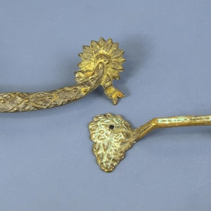 May include: Two brass drawer pulls with a weathered finish. The top pull features a curved, textured bar with floral rosettes at each end. The bottom pull has a straight, angular bar connecting two textured, leaf-shaped mounting plates.
