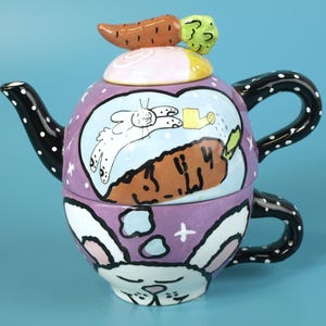 May include: A whimsical ceramic teapot and teacup set with a bunny theme. The teapot is lavender with a bunny illustration and a carrot-shaped lid. The teacup features a bunny face design. The handle and spout are black with white polka dots.