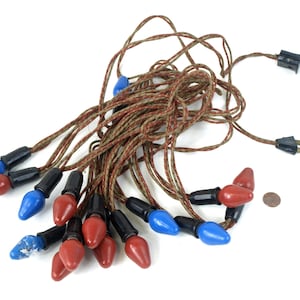 Puede incluir: Cadena de luces navideñas vintage con bombillas rojas y azules, bases negras y cable marrón y verde. Las luces tienen una forma clásica de lágrima y están dispuestas sobre un fondo blanco.