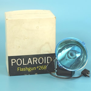 May include: Vintage Polaroid Flashgun #268 with a blue reflector and a black base. The flashgun is connected to a black cord and plug. The box is off-white with the Polaroid logo and Flashgun model number printed on it.