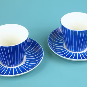 May include: Two matching blue and white striped coffee cups and saucers. The cups have a white interior and a blue handle. The saucers have a white rim and a blue striped design. The background is a solid light blue.