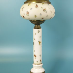 Könnte beinhalten: Eine antike Tischlampe mit weißem Glasschirm und Säule, verziert mit bunten Blumenmustern. Die Lampe hat Messingakzente, einen dekorativen Sockel und eine kreisförmige Oberseite. Die Lampe ist an eine Steckdose angeschlossen.