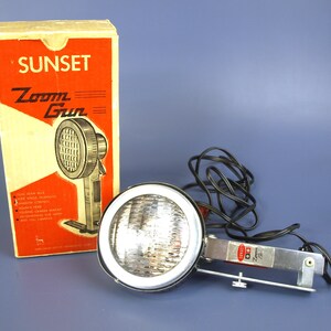 May include: Vintage Sunset Zoom Gun flash unit with original box. The flash has a round, chrome-rimmed light and a silver metal bracket. The box is cream and red with the words "Sunset Zoom Gun" printed on it.