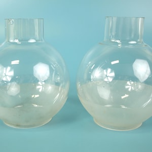 May include: Two clear glass lamp shades with a spherical body and a cylindrical neck. The lower portion of each globe features a frosted glass finish, while the upper section remains transparent, creating a distinct contrast in texture and clarity.