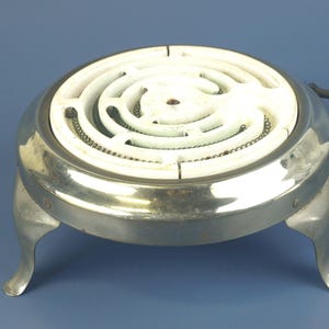 May include: A vintage electric stove burner with a white heating element and a silver-colored metal base. The burner has a circular design with a spiral heating coil and rests on three curved legs. A black power cord extends from the side.