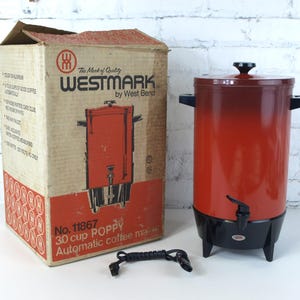 Könnte beinhalten: Vintage Westmark Poppy 30-Tassen-Kaffeeautomat mit Originalverpackung. Der Kaffeeautomat ist rot und schwarz mit einem Auslauf und Griffen. Die Box ist beige mit rotem und schwarzem Text und Grafiken.