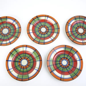 Może przedstawiać: Pięć ceramicznych talerzy z kolorowym wzorem w kratę. Każdy talerz ma koncentryczne okręgi w kolorze białym, czerwonym, zielonym i niebieskim, obramowane pomarańczową obwódką. Talerze ułożone są na białej powierzchni.
