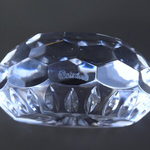 May include: A clear, faceted crystal object with a geometric design. The object has a rounded shape with a series of hexagonal and leaf-shaped cut patterns. The word "Waterford" is etched on the surface.