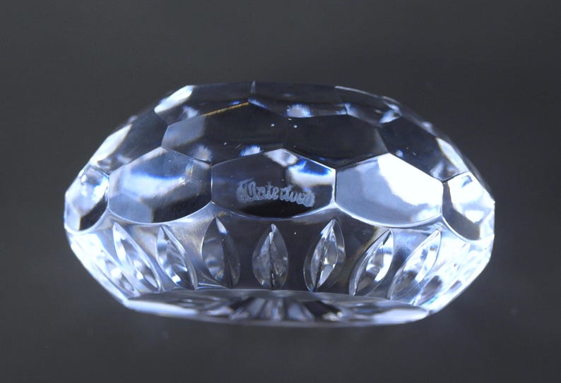 May include: A clear, faceted crystal object with a geometric design. The object has a rounded shape with a series of hexagonal and leaf-shaped cut patterns. The word "Waterford" is etched on the surface.
