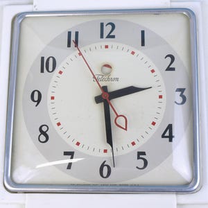 以下が含まれることがあります： 白文字盤、黒数字、赤のアクセントが特徴のヴィンテージTelechron時計。銀色のフレームと白いプラスチックケースを備えた正方形の形状です。針は黒と赤です。時計は約2時55分を示しています。