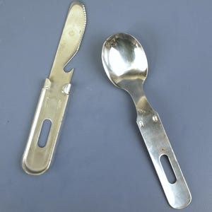 Op de afbeelding: Een zilverkleurige kampeerbestekset, bestaande uit een mes met een gekartelde rand en flesopener, en een lepel. Beide gebruiksvoorwerpen hebben een handvat met een gat om op te hangen. De set is ontworpen voor gebruik buitenshuis.
