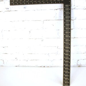 May include: A black metal framing square featuring white numerical markings and measurement scales along both edges. The tool displays metric units with clear increments, including fractional markings and various conversion tables printed on the surface.
