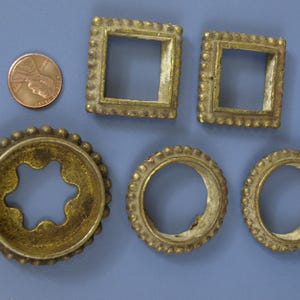 May include: An assortment of antique brass metal shapes, including two square frames, three circular frames, and a star-shaped frame. A penny is included for size comparison. The objects have a tarnished, aged appearance.