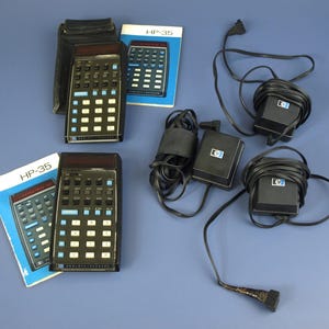 Könnte beinhalten: Vintage HP-35-Taschenrechner mit schwarzen Gehäusen und weißen und blauen Tasten. Das Bild enthält zwei Taschenrechner, zwei Handbücher und mehrere schwarze Netzadapter. Die Handbücher sind mit "HP-35" beschriftet. Die Taschenrechner befinden sich auf einer blauen Oberfläche.