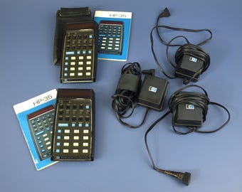 Puede incluir: Calculadoras HP-35 vintage con carcasas negras y teclas blancas y azules. La imagen incluye dos calculadoras, dos manuales y varios adaptadores de corriente negros. Los manuales están etiquetados como "HP-35". Las calculadoras están sobre una superficie azul.