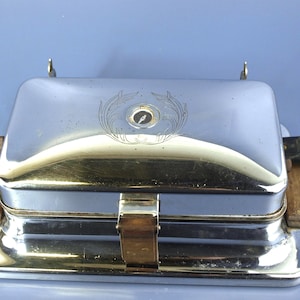 May include: A vintage chrome waffle iron with wooden handles and a decorative emblem on the lid. The appliance has a rectangular shape and a black power cord. The waffle iron is a classic kitchen appliance.