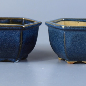 Puede incluir: Dos macetas de cerámica hexagonales con un acabado de esmalte azul profundo y moteado. Cada maceta presenta un interior liso de color claro y un borde de tono metálico oscuro. Las macetas se apoyan sobre pequeños pies integrados en la base.
