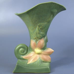 May include: A green ceramic vase with a flared top and a floral design. The vase features a large, stylized flower in shades of pink and yellow, with green leaves and a decorative swirl on the side. The vase is approximately 10 inches tall.