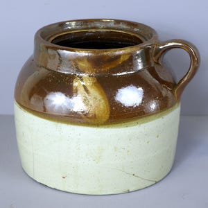 Puede incluir: Un tarro de barro cocido esmaltado en marrón y blanco con una sola asa. El tarro tiene una boca ancha y un cuerpo redondeado.
