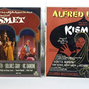 Op de afbeelding: Twee vintage vinyl platenhoezen voor de musical Kismet. De linker hoes heeft een kleurenfoto van de cast voor een rode en gouden achtergrond. De rechter hoes heeft een zwart-witte illustratie van een man in een tulband en een gewaad. Beide hoezen hebben de titel Kismet in gouden letters.