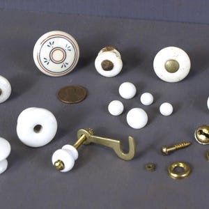 May include: Assortment of white ceramic knobs with a variety of designs, including a floral pattern, a plain round knob, and a knob with a raised centre. There are also several small white ceramic beads and a brass hook.