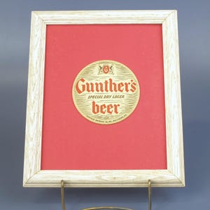 May include: Framed vintage Gunther's Beer advertisement. The circular emblem, set against a red background, features the brand name and "Special Dry Lager" text. The frame is a light wood color, and the piece is displayed on a metal stand.
