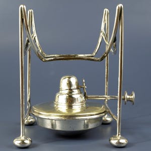 Puede incluir: Un calienta platos plateado con un soporte. El soporte tiene cuatro patas con pies redondeados y una parte superior curvada para sostener un plato. El plato tiene un quemador con una perilla y una tapa pequeña en forma de cúpula. El calienta platos está diseñado para mantener la comida caliente.