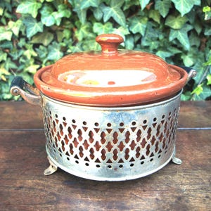 May include: A vintage terracotta casserole dish with a lid, set in a decorative silver metal stand. The dish has a round shape with a small handle on the lid. The stand has a pierced design and three small feet.