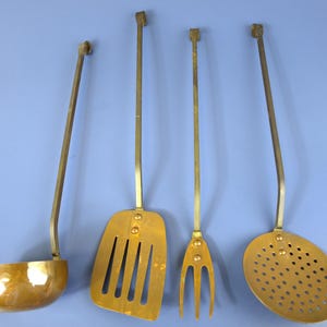 May include: A set of four brass kitchen utensils featuring long, slender handles with square-shaped ends. The collection includes a deep round ladle, a slotted spatula, a three-pronged fork, and a circular skimmer with numerous small perforations.