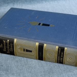May include: A blue hardcover book titled "The Illustrated Encyclopedia Library" with gold lettering and a gold and black spine. The book is a Standard Family Cook Book.